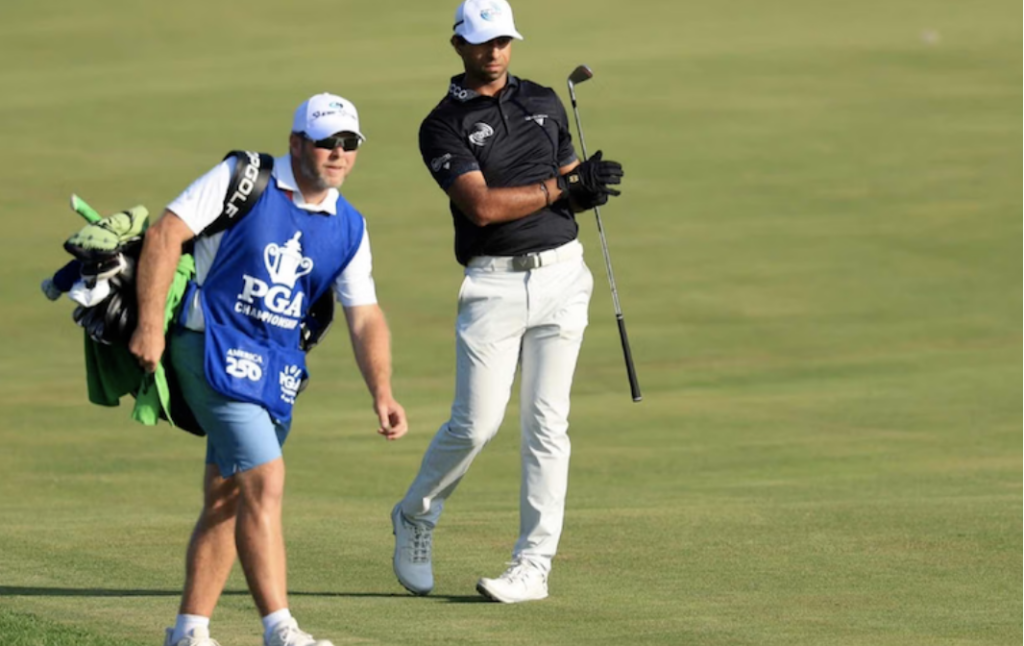 Two golfers on a green golf course: a caddie in a blue PGA vest carrying clubs walks beside a pro in a black polo preparing to swing with a golf club