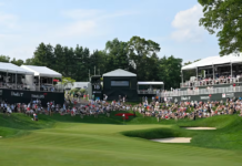 The Travelers Championship Live Leaderboard Crowd watching a golf tournament from grandstands along a green fairway on a sunny day, with sand bunkers nearby and a red flag hole in view.