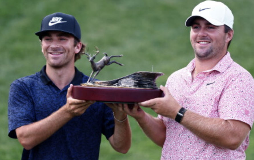 Two golfers in caps smile while presenting a trophy featuring a bird sculpture on a red base outdoors